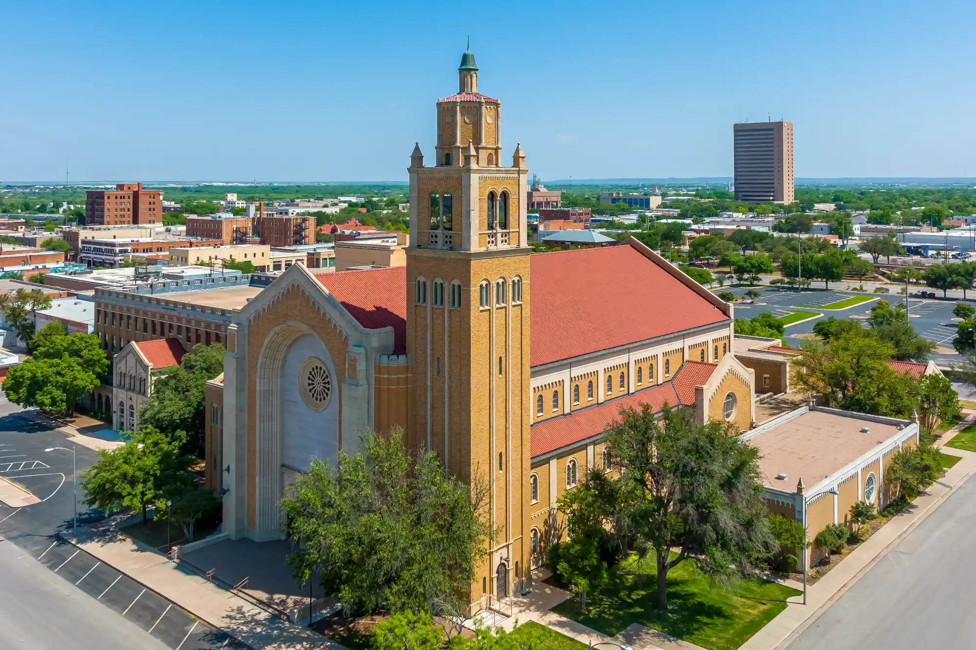 An aerial view of a tall, light tan church building with a red tile roof and a central bell tower, set among green trees in an urban skyline.