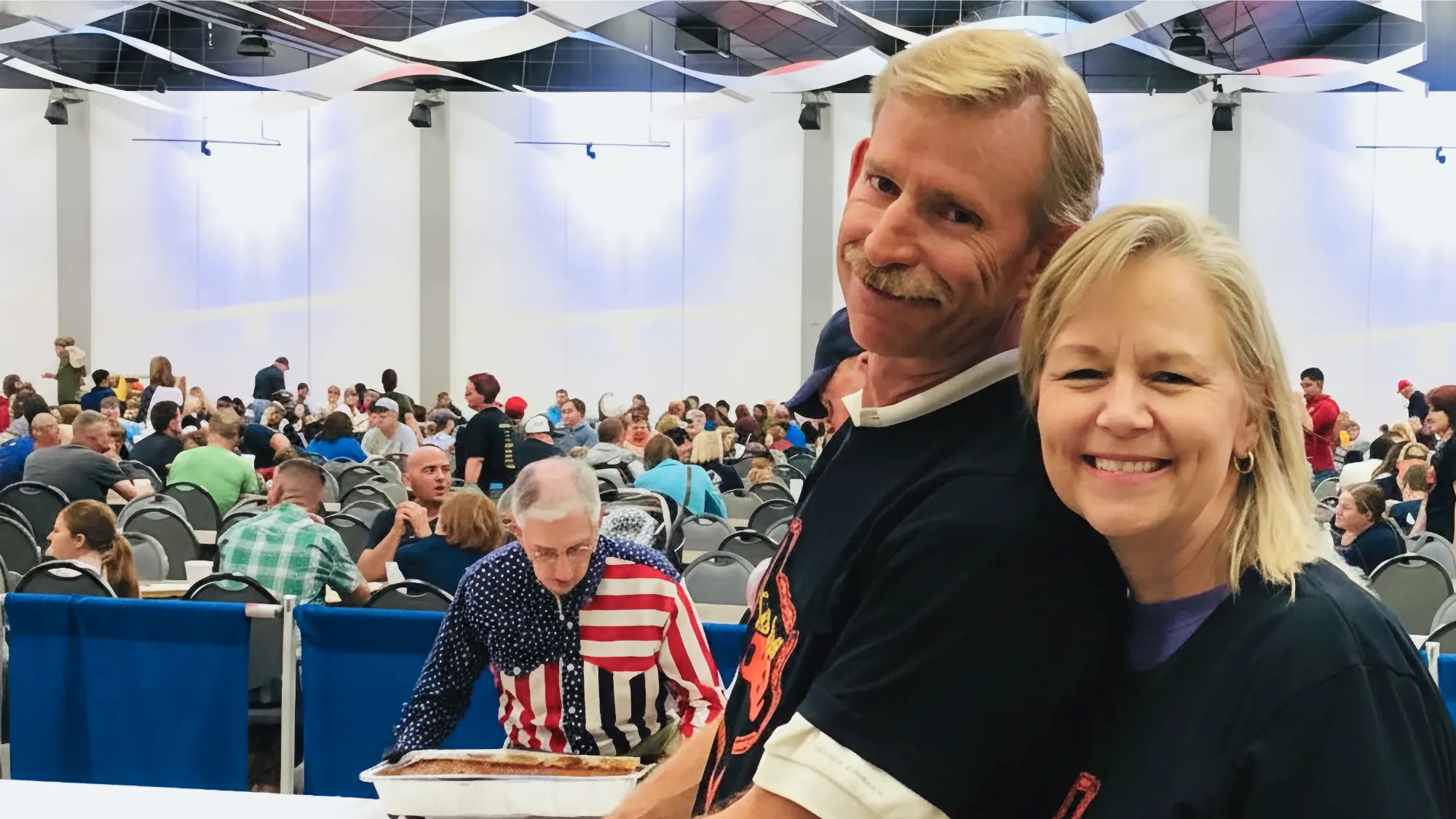 Chris Love, with a mustache, and Linda Love smile broadly in a large, busy hall where many people are seated at tables.
