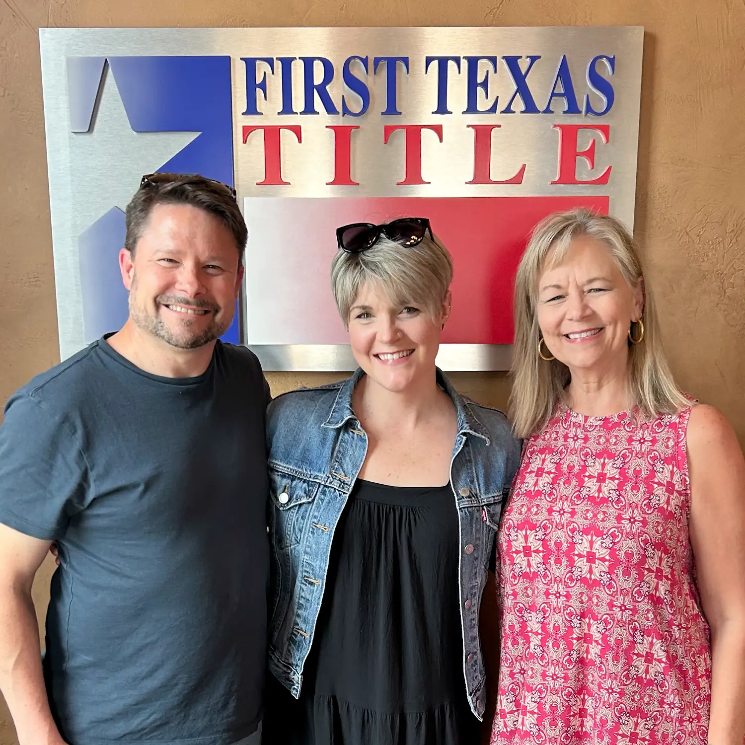 Three smiling people stand together in front of a First Texas Title sign. The man wears a blue t-shirt, the center woman a denim jacket, and the woman on the right wears a pink patterned dress.