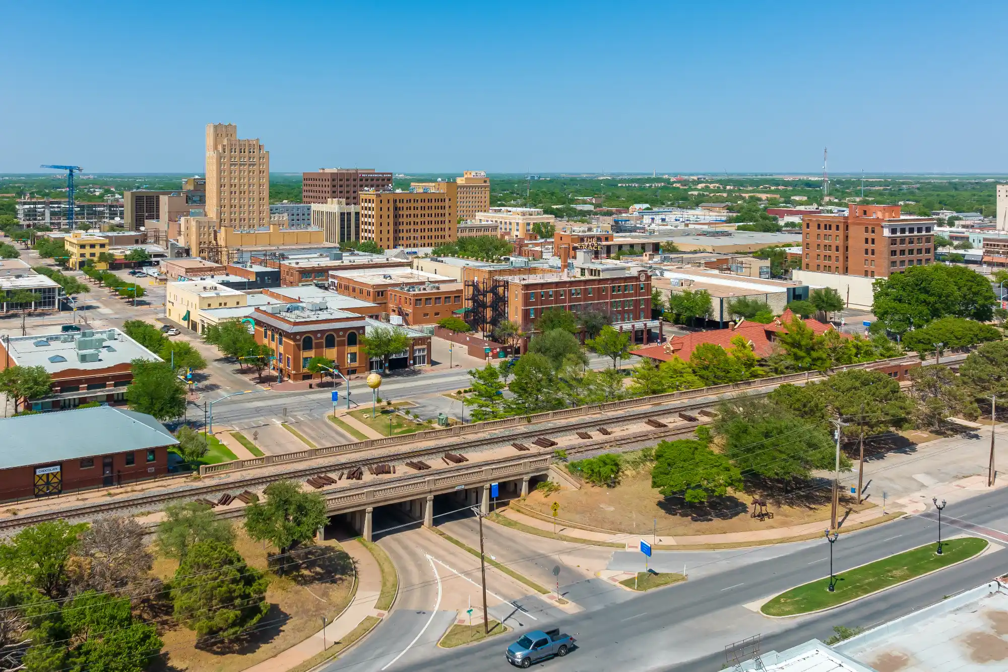 An aerial view of a sunny downtown cityscape featuring historic and modern buildings. A prominent elevated railway overpass crosses the foreground above city streets.