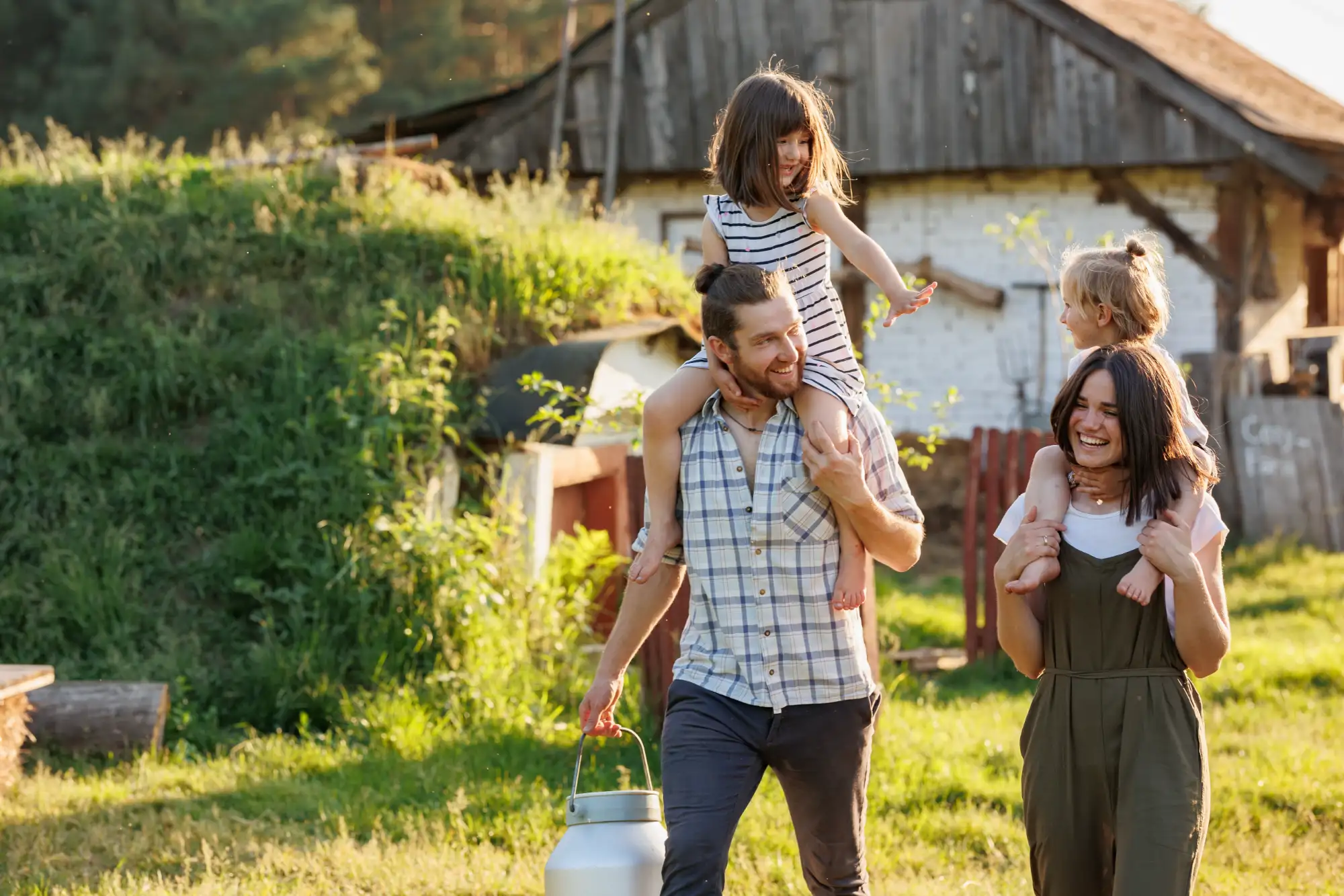 A father and mother, both smiling, carry their two young daughters on their shoulders through a grassy farmyard. The father holds a metal milk can.