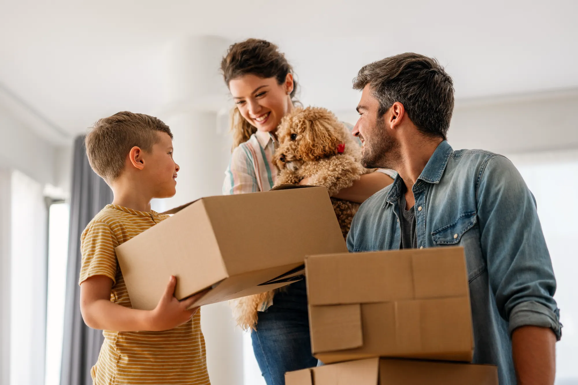 A smiling family, including a father, mother holding a dog, and their son, happily carry moving boxes into their bright, new home.