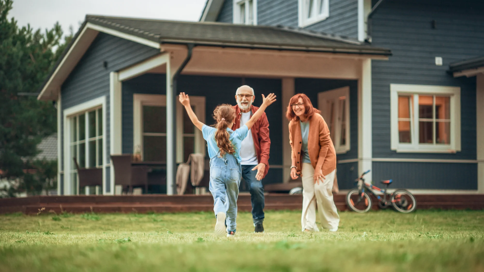 A little girl in overalls runs across the lawn toward her smiling grandmother and grandfather standing on their home's porch.