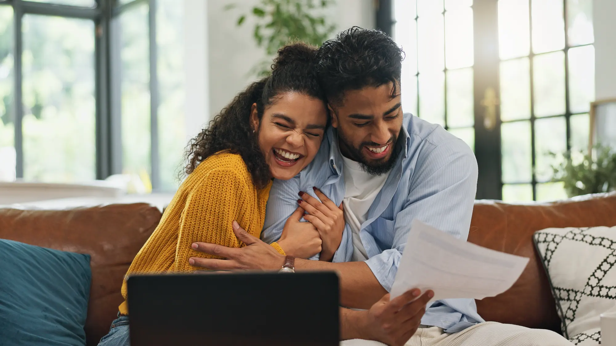 A laughing couple sits close on a leather couch; the man holds a document while the woman, wearing a yellow sweater, hugs him, celebrating good financial news.