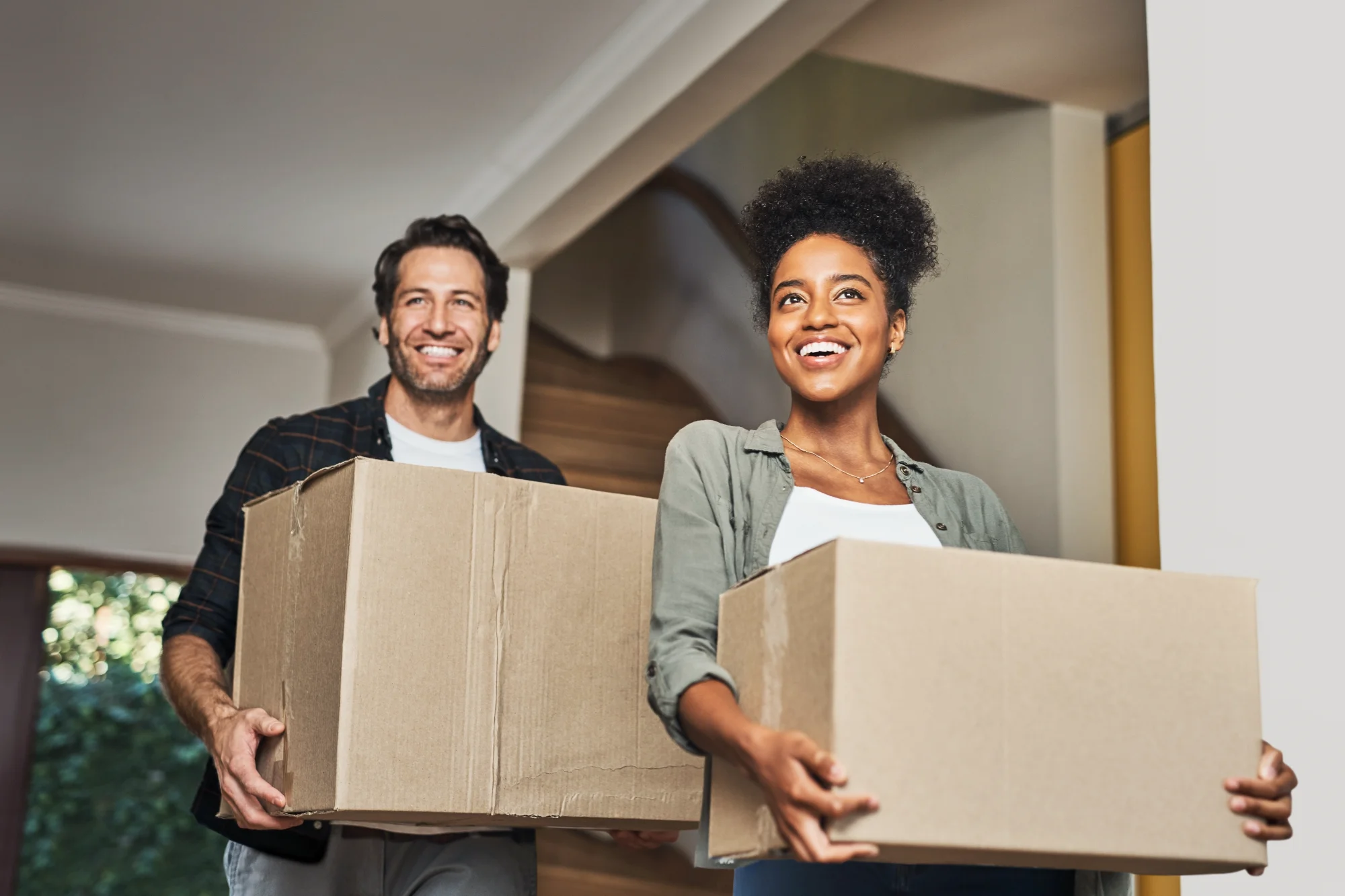 A happy couple smiles brightly as they carry moving boxes through the foyer of their new home.