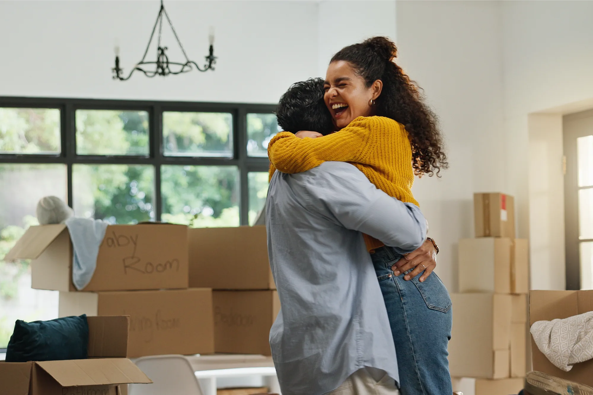 A couple embraces in a bright room filled with moving boxes, one labeled "Baby Room." The woman is laughing with an expression of pure happiness.