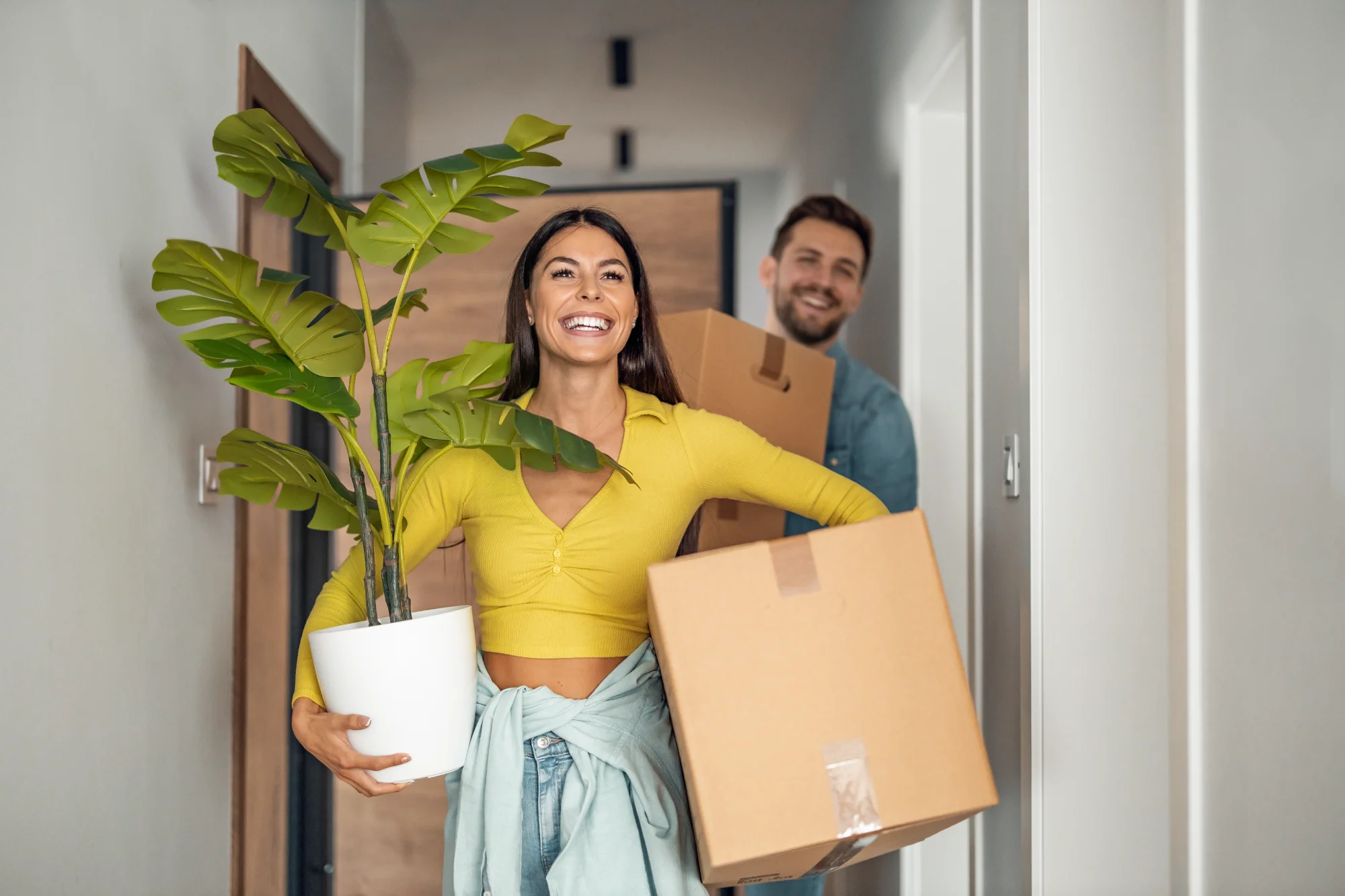 A woman in a yellow top, smiling brightly, carries a potted plant and a box into a home, followed by a smiling man carrying another box.