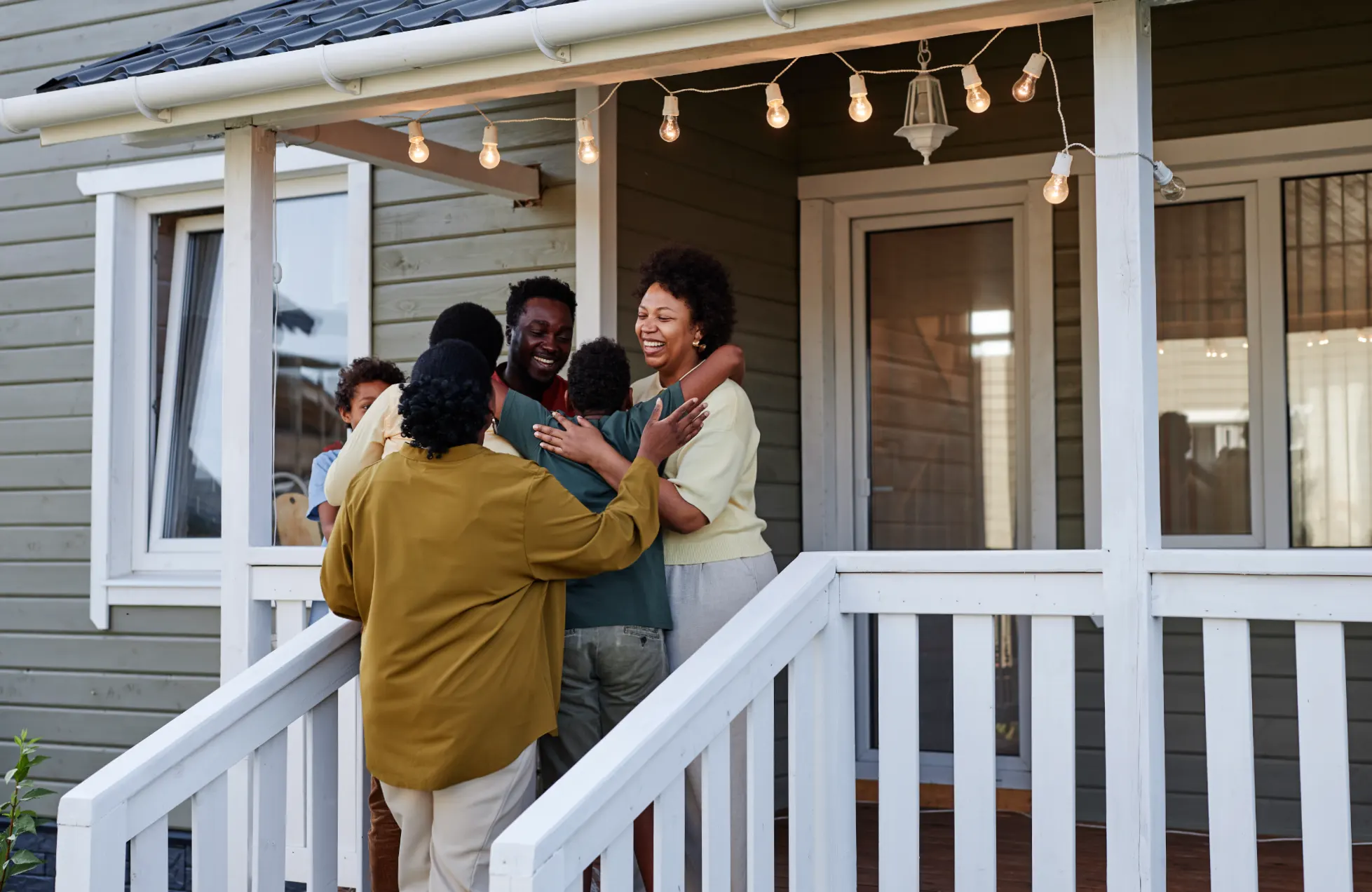 A happy family, including children and adults, shares a heartfelt group hug on the front porch of a house with white railings.