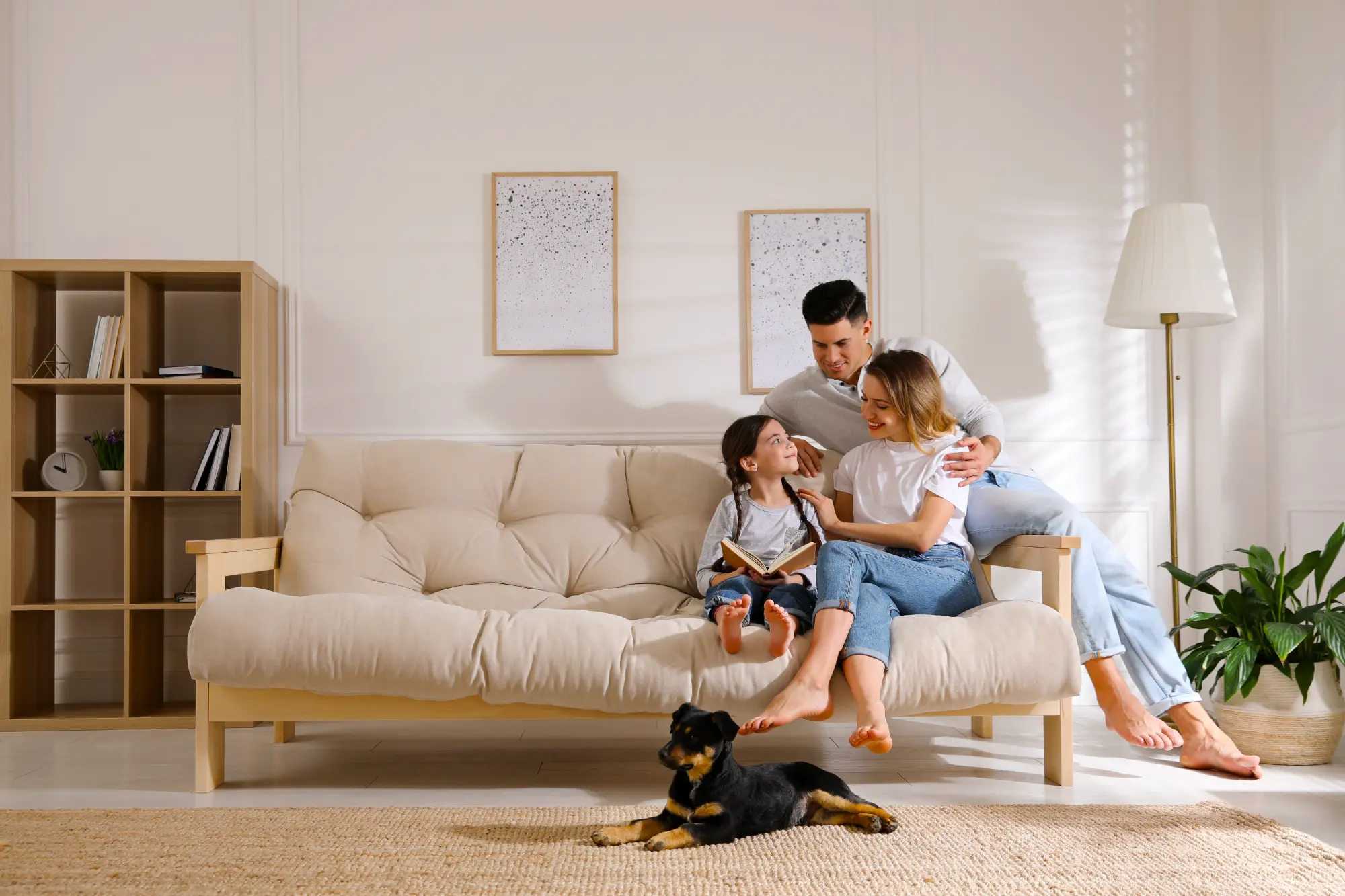 A mother, father, and daughter smile happily on a couch in a living room, while a black and tan dog rests on the rug.