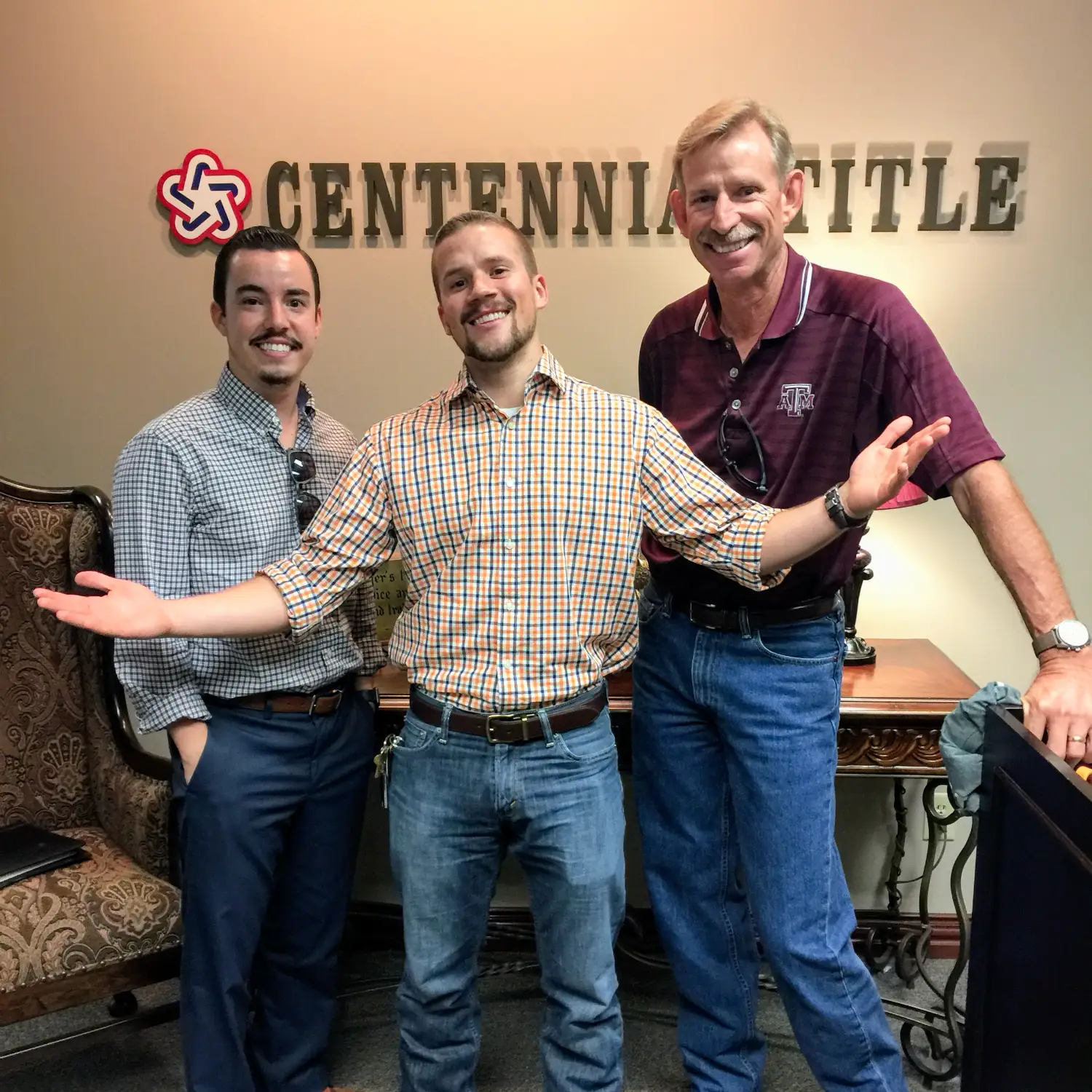 Three men stand in an office in front of the "Centennial Title" sign, all smiling happily. One man with a plaid shirt extends his arms in a welcoming gesture.
