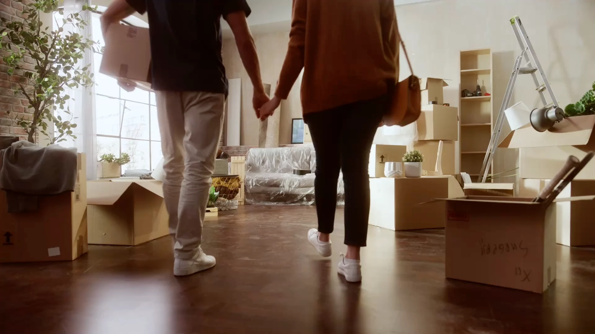 A man carrying a box and a woman walk hand-in-hand through a brightly lit room filled with numerous moving boxes and plastic-covered furniture.