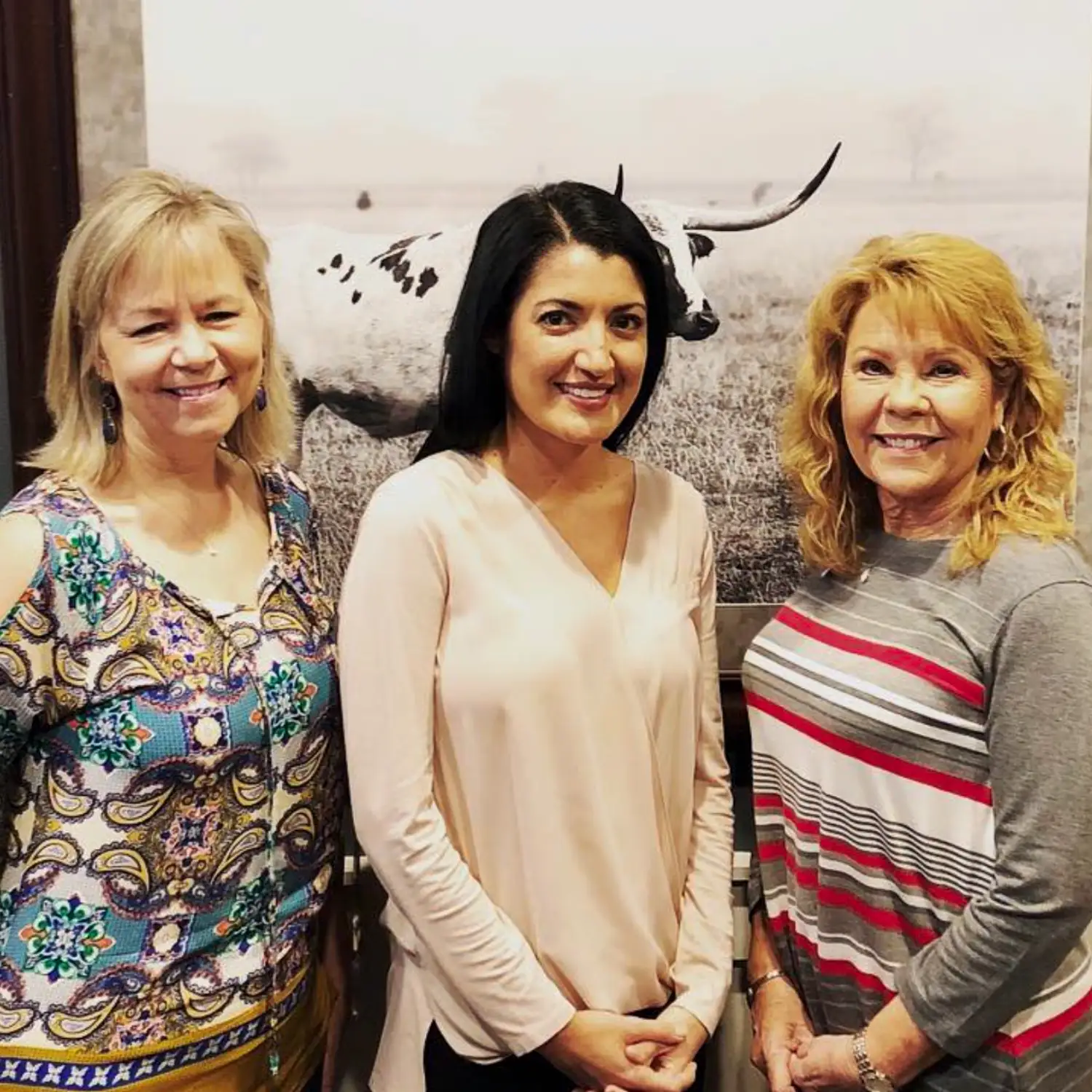 Three smiling women pose together, celebrating a successful home closing. They stand indoors in front of a large black and white image of a Longhorn steer.