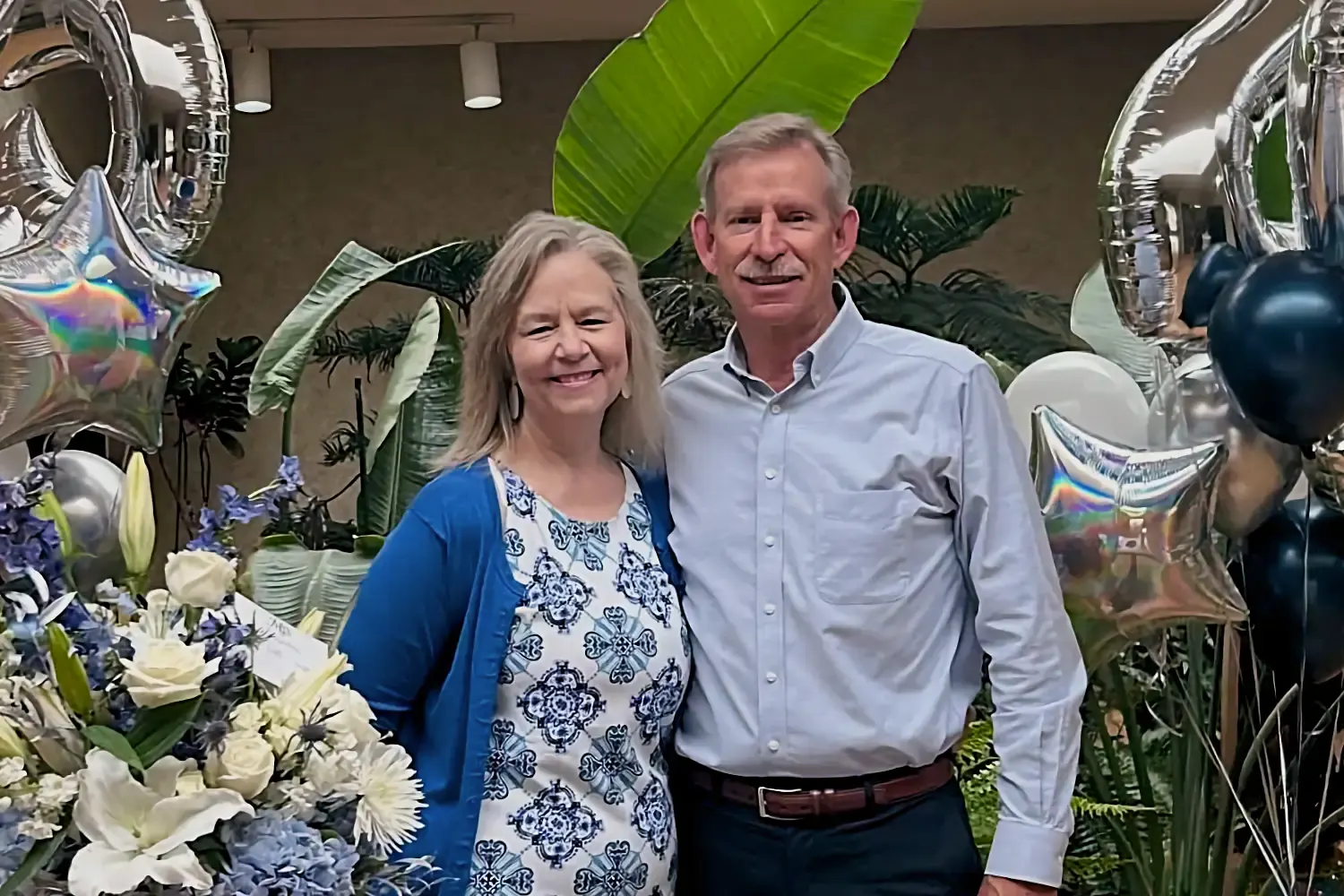 Linda and Chris, smiling warmly, stand together at their 25th-anniversary party, surrounded by large plants and festive silver and blue decorations.