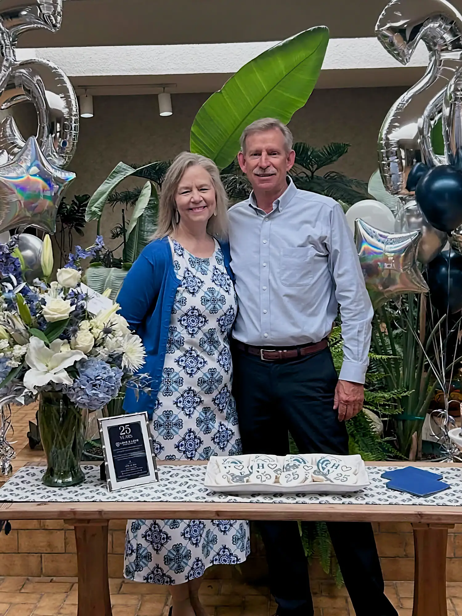A couple, Linda and Chris Love, smile proudly at their 25th anniversary celebration, with balloons, flowers, and a sign reading "25 Years Love & Love."