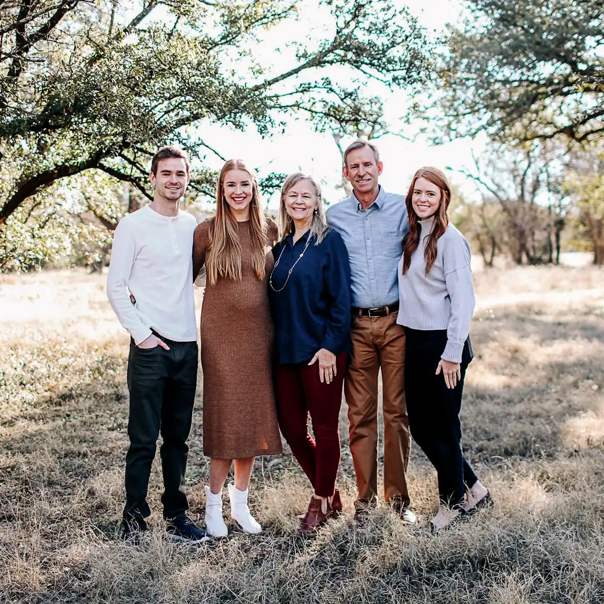 A happy family of five, comprised of two parents and three adult children, smiling together for a portrait in a sunny outdoor setting.