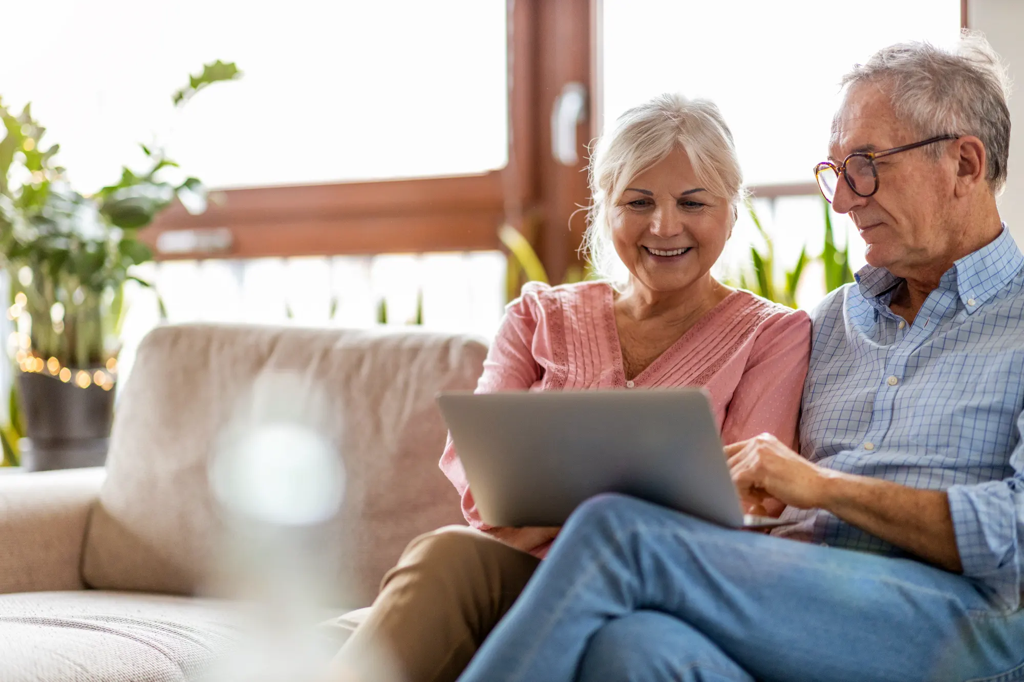 A smiling older couple sits on a couch, intently browsing content on a shared laptop. The man in glasses is pointing at the screen as the woman watches happily.