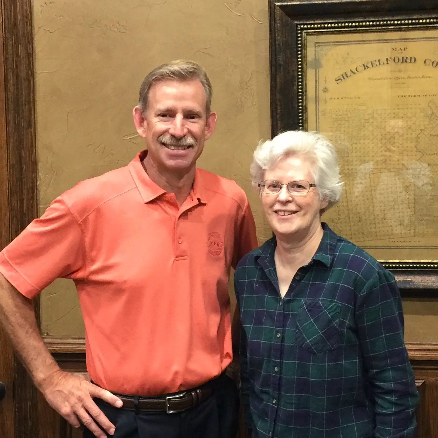 A man in an orange polo shirt with a mustache stands next to an older woman in glasses and a plaid shirt. Both are smiling warmly.