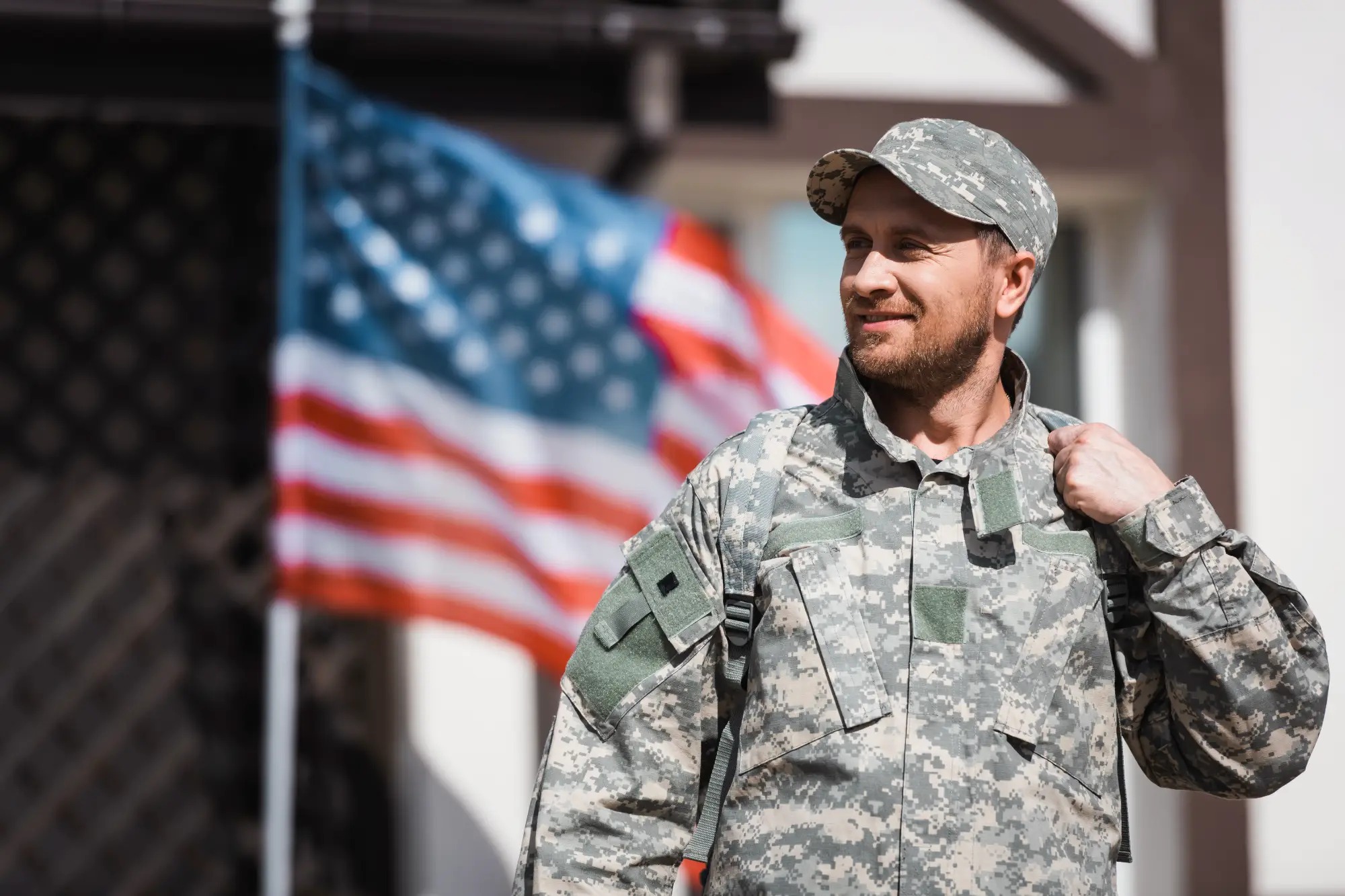 A veteran in digital camouflage fatigues and a cap smiles with a positive expression while standing in front of a blurred American flag.