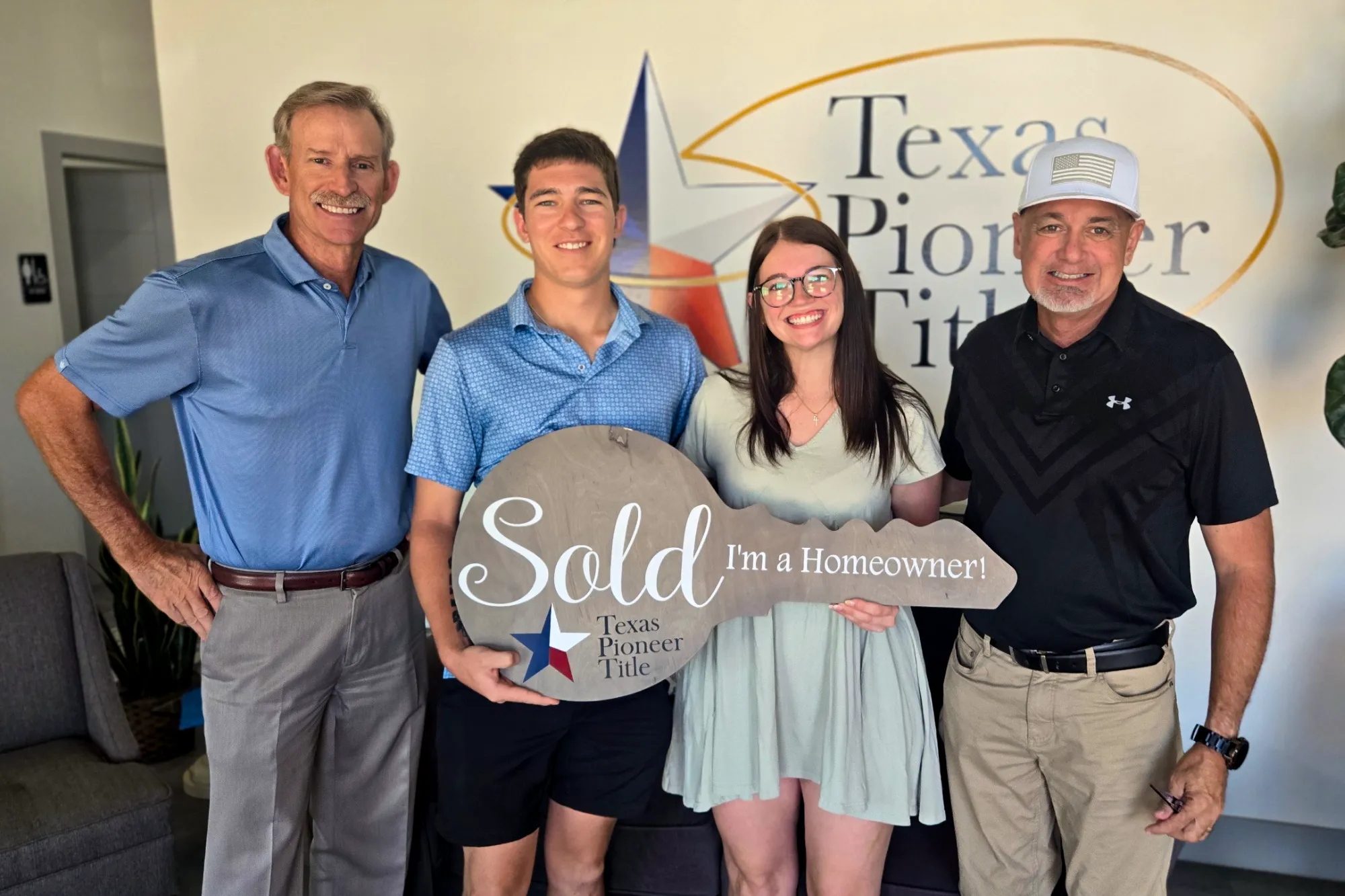 Two men stand with a joyful young couple celebrating the purchase of their first home. The couple holds a key-shaped sign reading, "Sold I'm a Homeowner!"